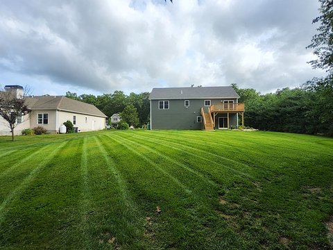 Large backyard lawn mowed in diagonal light and dark green stripes, with a house and wooden deck at the back under an overcast sky
