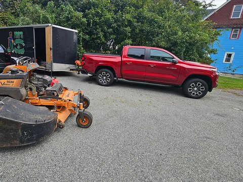 Red crew-cab pickup hitched to a black enclosed trailer with the Fro's Landscape and Construction logo, and an orange Scag stand-on mower in front